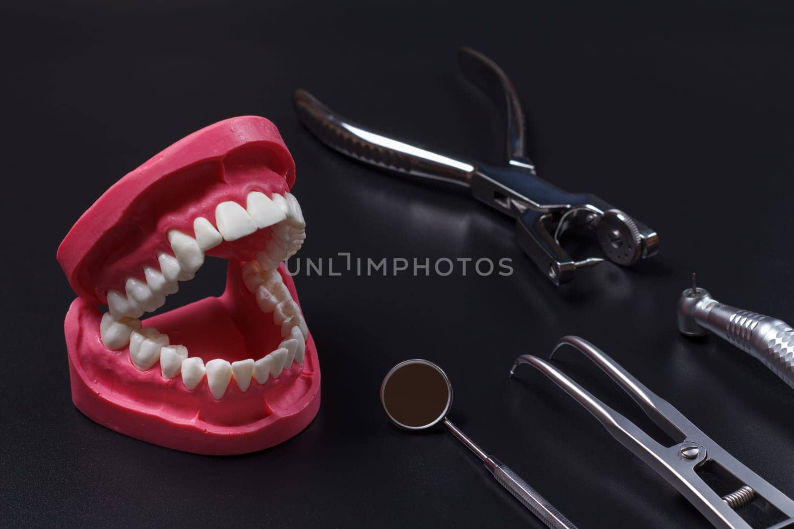 Layout of a human jaw with the rubber dam forcep and the dental hole punch. Head of high-speed dental handpiece with bur, a metal mirror on the black background.