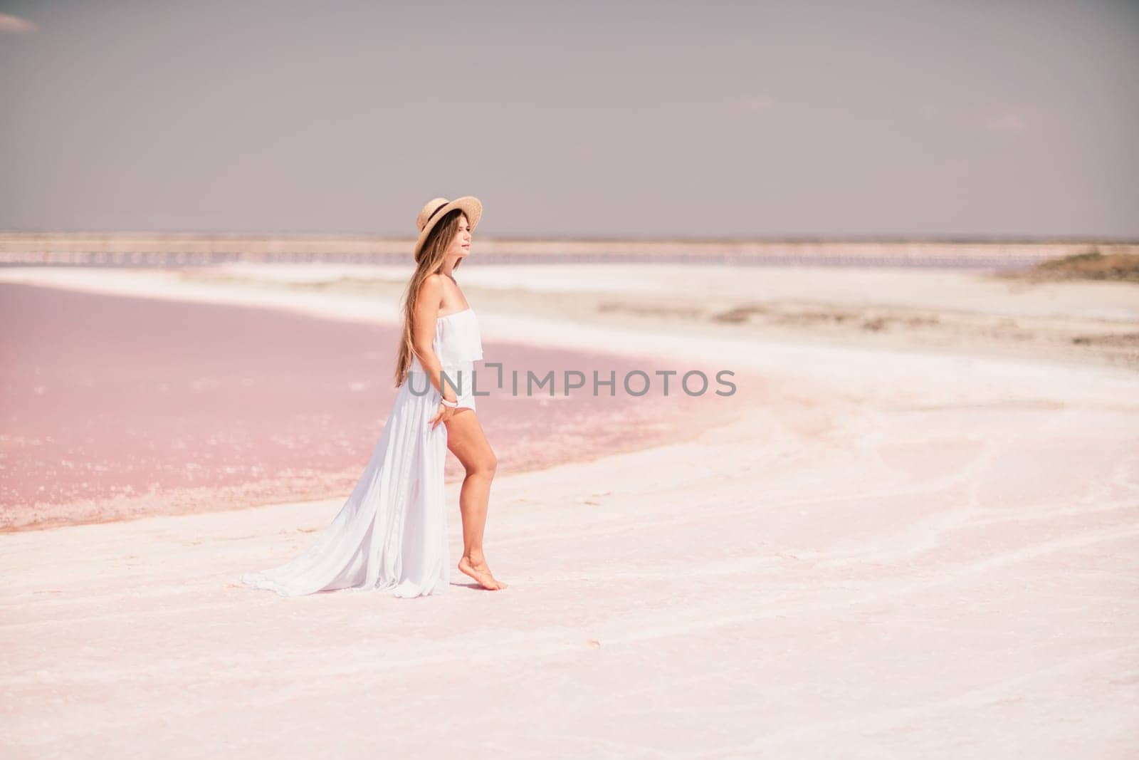 Woman in pink salt lake. She in a white dress and hat enjoys the scenic view of a pink salt lake as she walks along the white, salty shore, creating a lasting memory