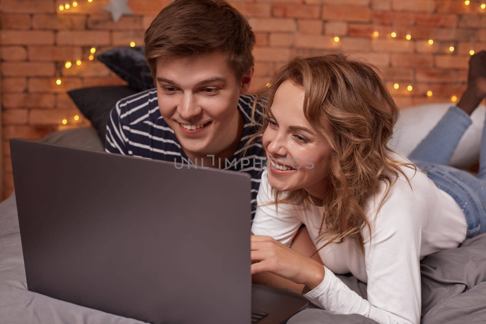 Smiling young man and woman lying in bed and watching something on the laptop. They're happy and lovely