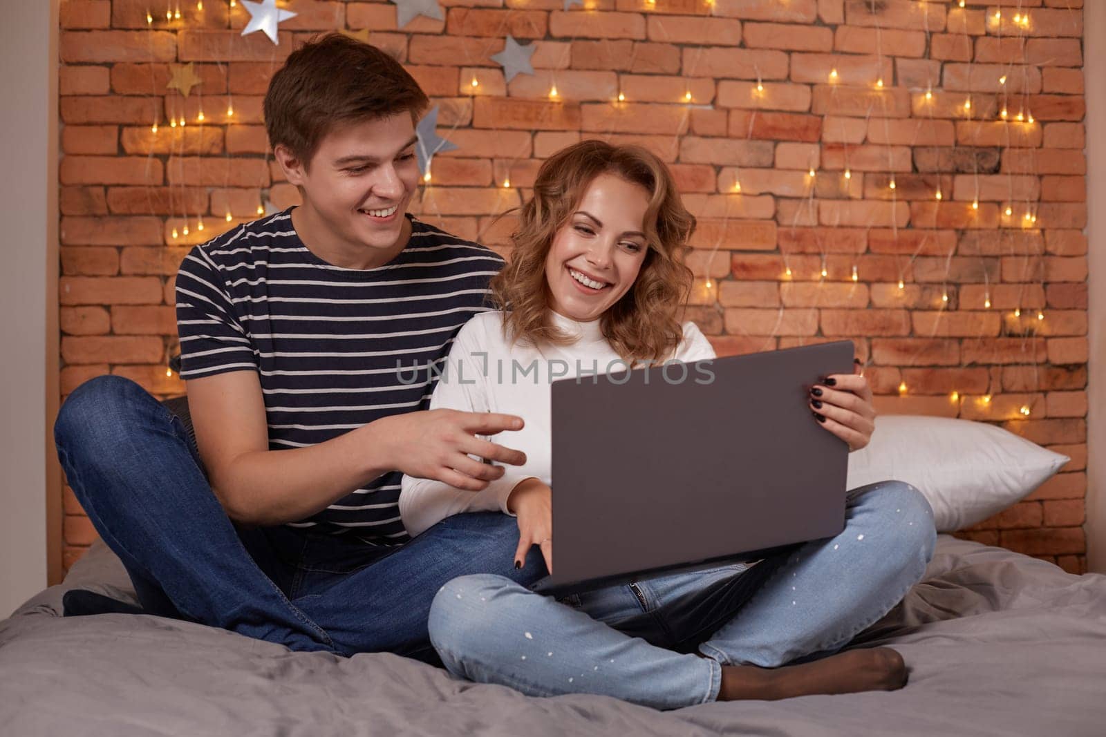 Smiling young couple man and woman sitting on bed and watching something on the laptop. They have fun together