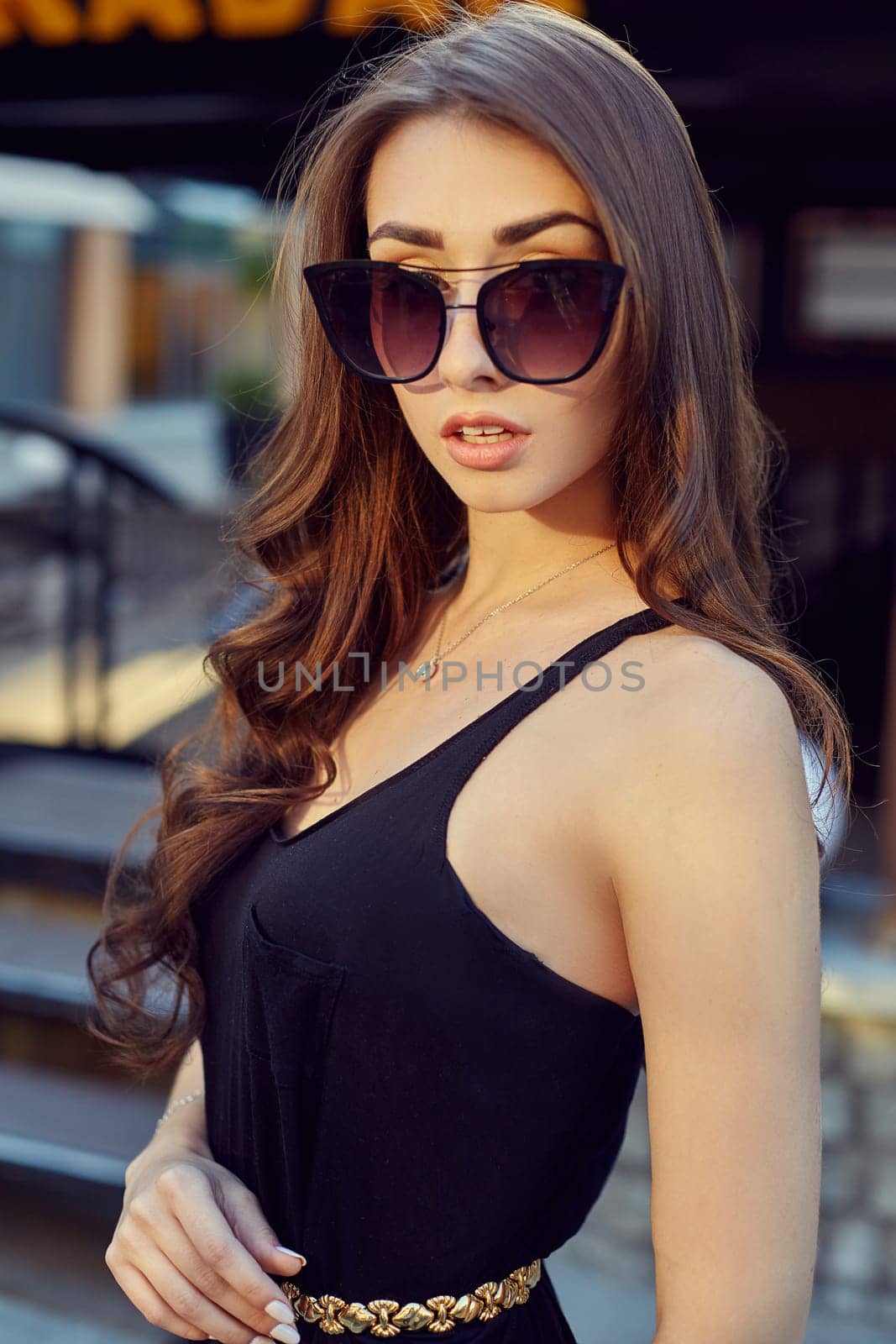 Fashion style portrait of young beautiful elegant woman in black dress, sunglasses, with long dark hair walking at city streets.