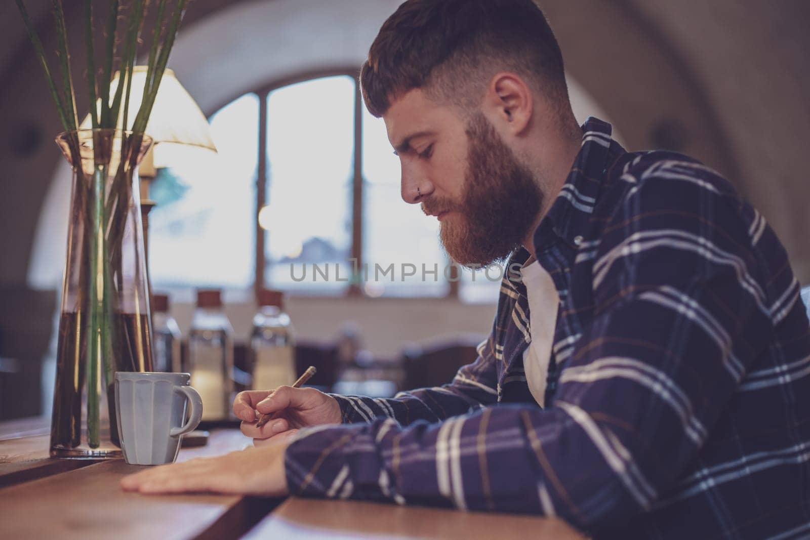 Young bearded businessman sits in cafe, home at table and writes by nazarovsergey