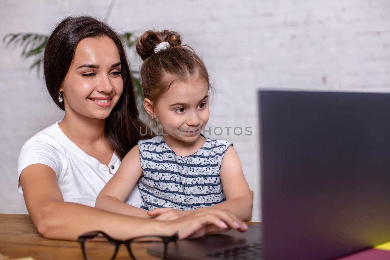 Attractive young woman and her little cute daughter are sitting at the table and having fun while doing homework together. Mother helps daughter with her school classes.
