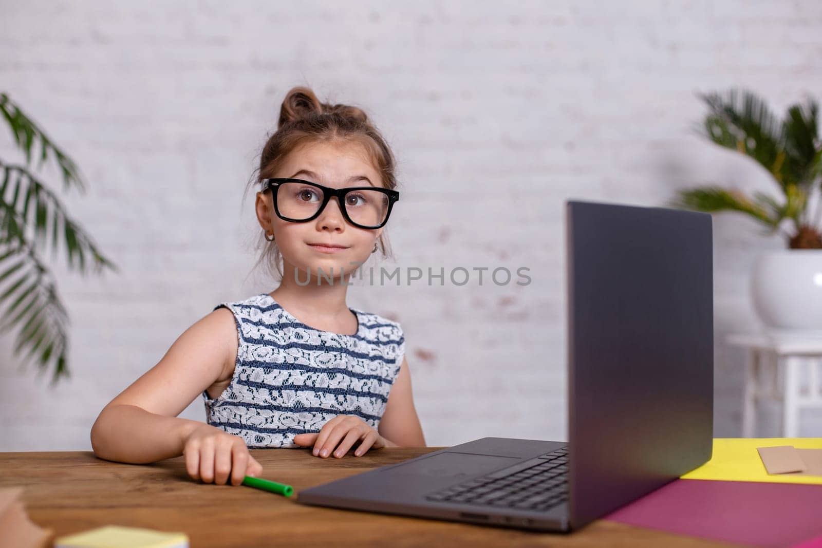 Cute little girl is sitting at table with her laptop and notebook, wearing glasses. The concept of new programs for teaching children