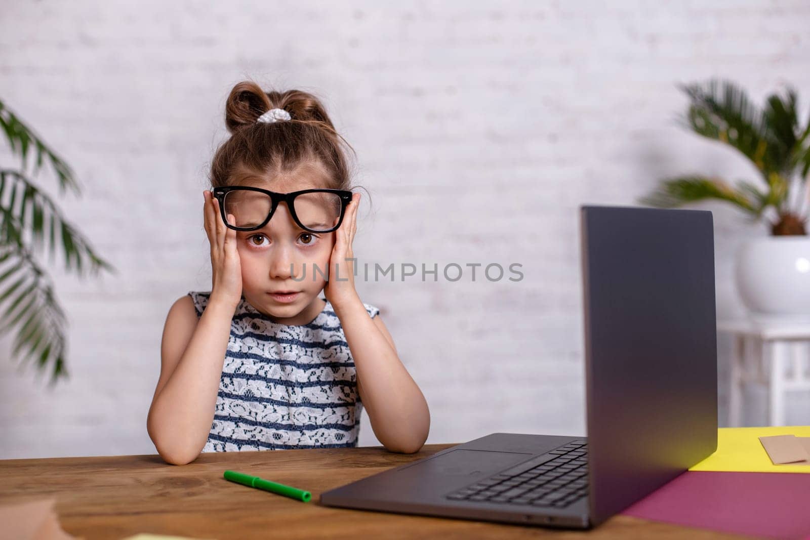 Cute little girl is sitting at table with her laptop and notebook, wearing glasses by nazarovsergey