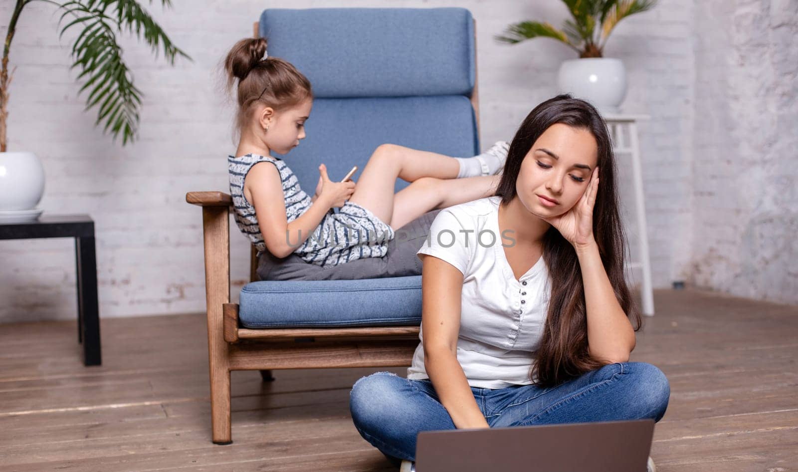 Mother working with laptop, sitting on the floor and cute little girl laying on the armchair and playing games on the smartphone