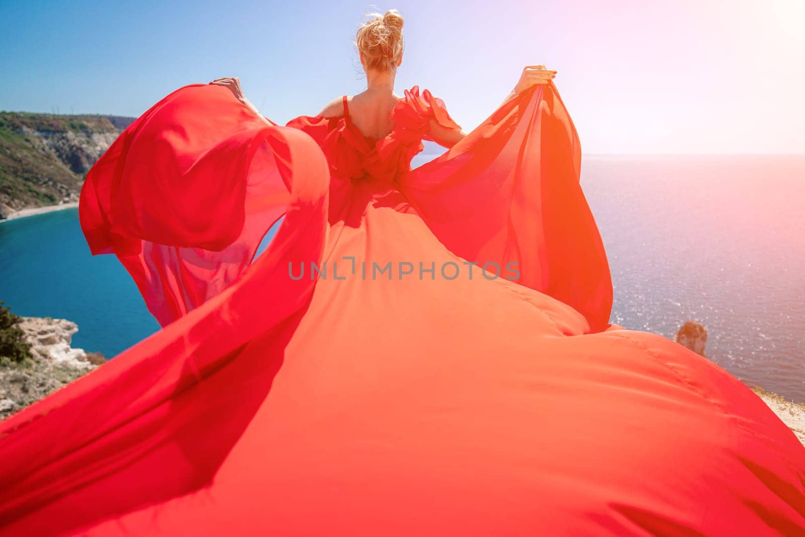 woman sea red dress. Blonde with long hair on a sunny seashore in a red flowing dress, back view, silk fabric waving in the wind. Against the backdrop of the blue sky and mountains on the seashore