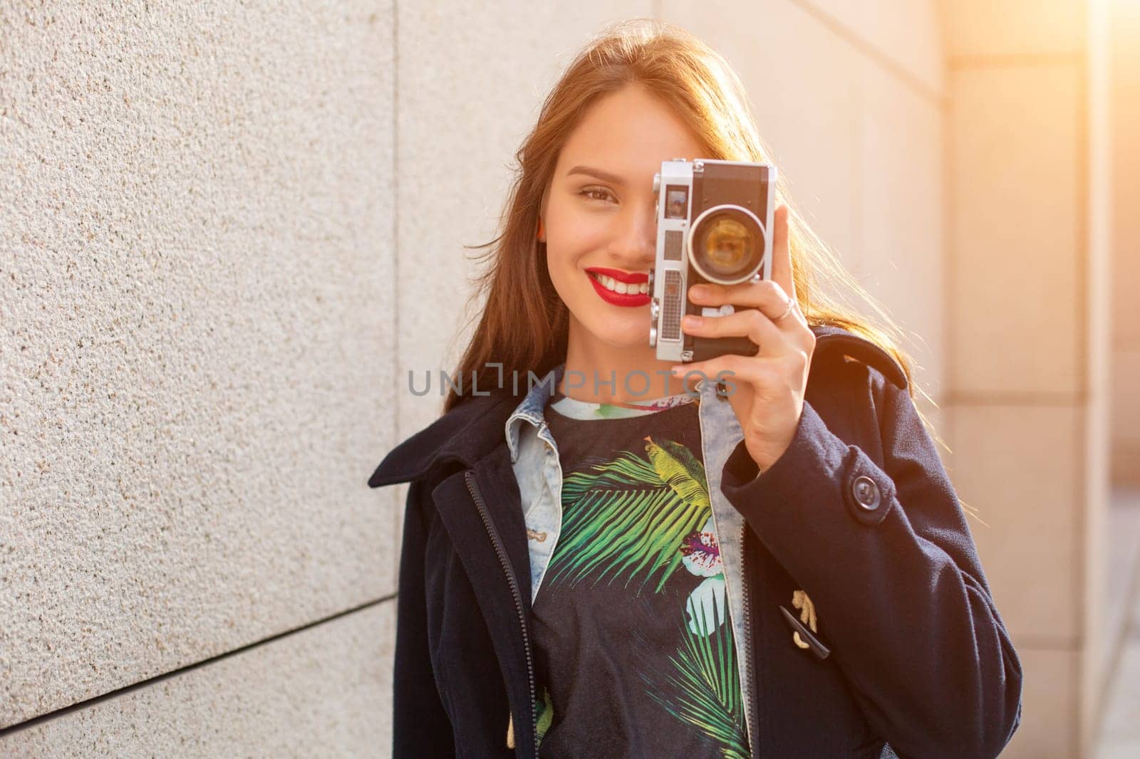 Happy hipster girl making photo with retro camera on city street. Sun flare by nazarovsergey