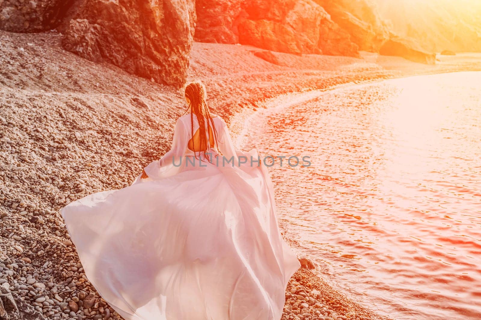 Mysterious woman silhouette long hair walks on the beach ocean water, sea nymph wind listens to the wave. Throws up a long white dress, a divine sunset. Artistic photo from the back without a face by Matiunina