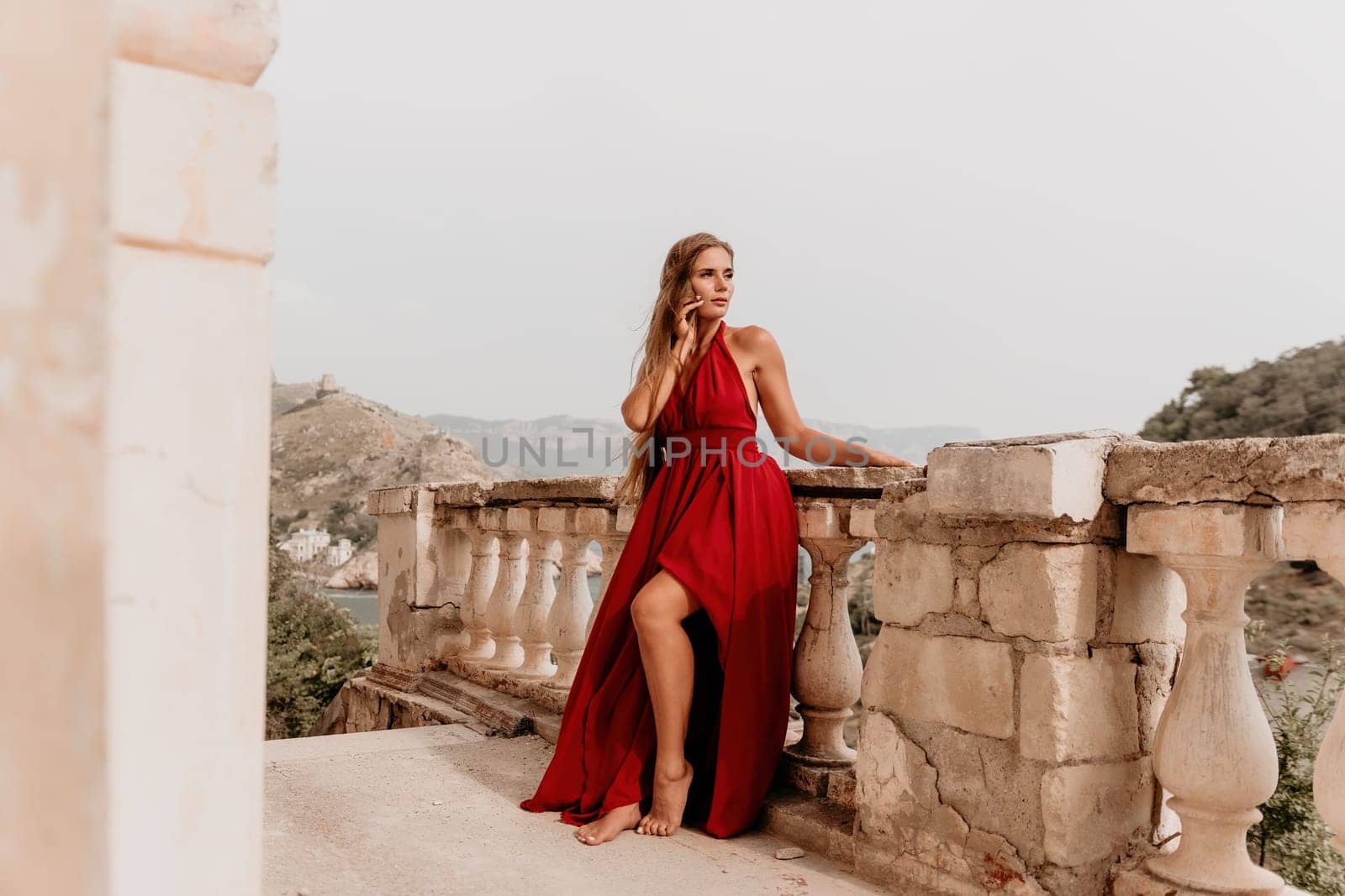 Side view a Young beautiful sensual woman in a red long dress posing on a volcanic rock high above the sea during sunset. Girl on the nature on overcast sky background. Fashion photo