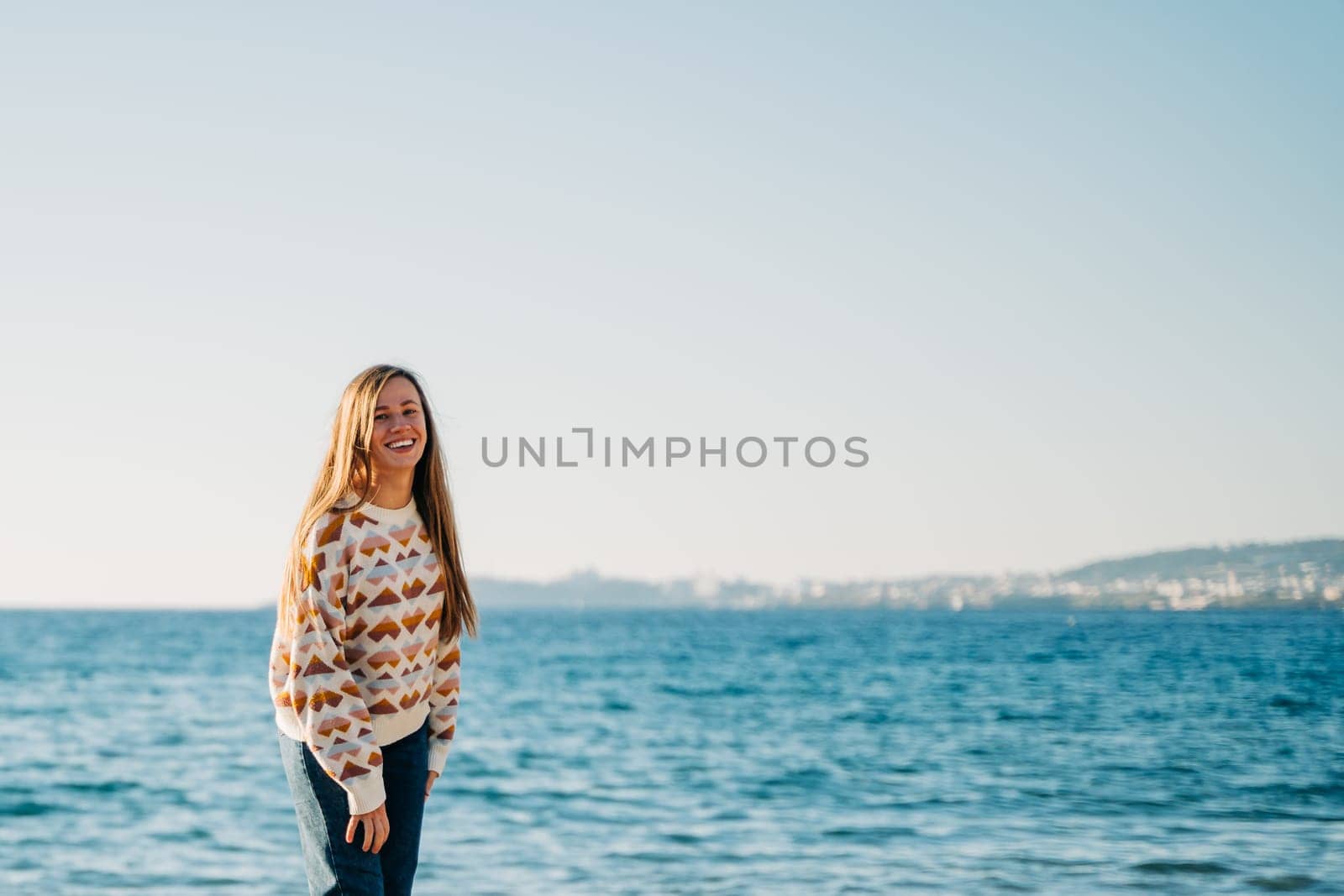 Portrait of Young beautiful woman in cozy sweater smiling and enjoying sunbathe on the sand beach near winter ocean. Cute attractive girl relaxing under the sun on autumn sea side shore.