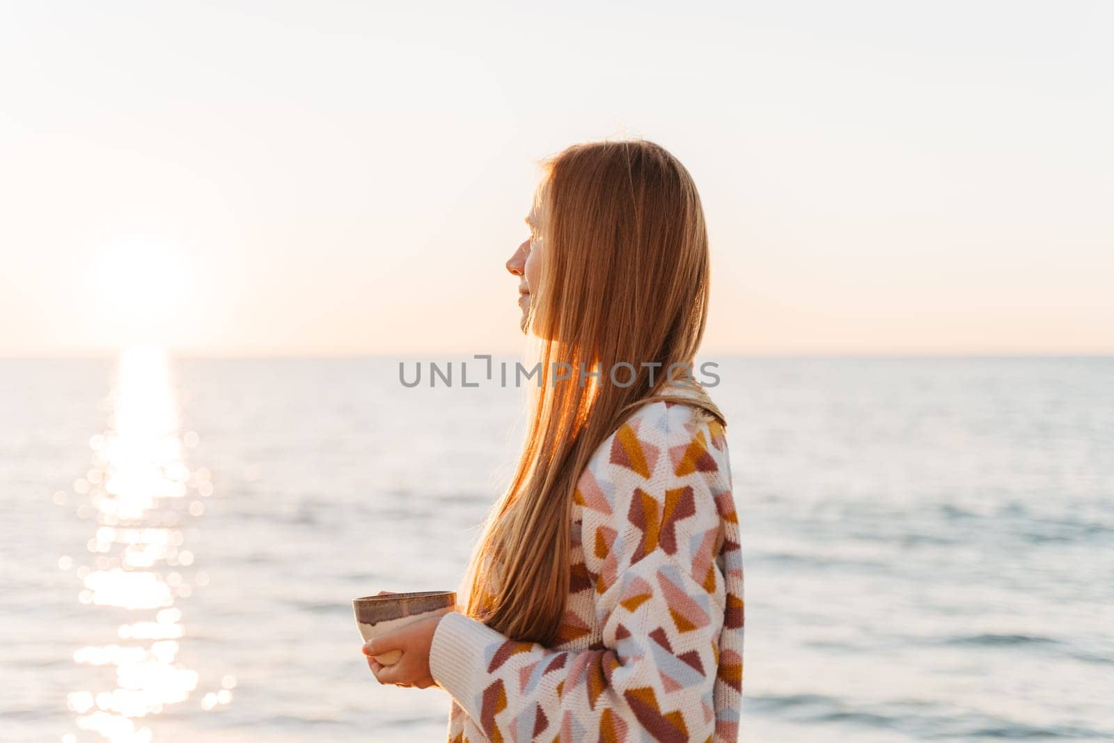 Side portrait of beautiful girl in cozy sweater holding a cup of coffee. Young attractive woman enjoying a view of winter seaside shore with sun trail on the surface of romantic autumn ocean by Ostanina