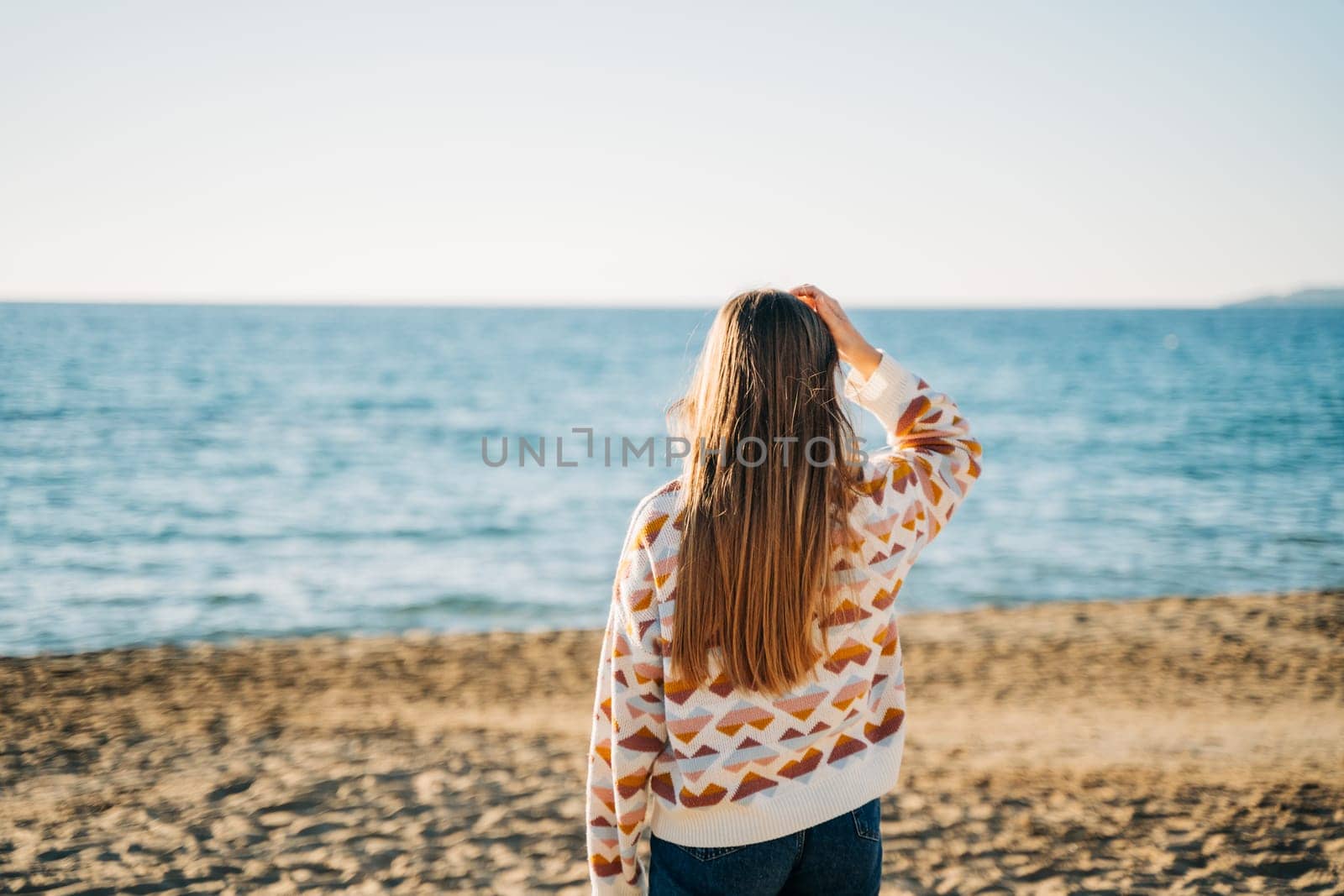 Back portrait of a girl in cozy sweater and jeans running on the winter beach to the sea. Woman walking on the autumn seashore sand and enjoying the view of a fall ocean waves.