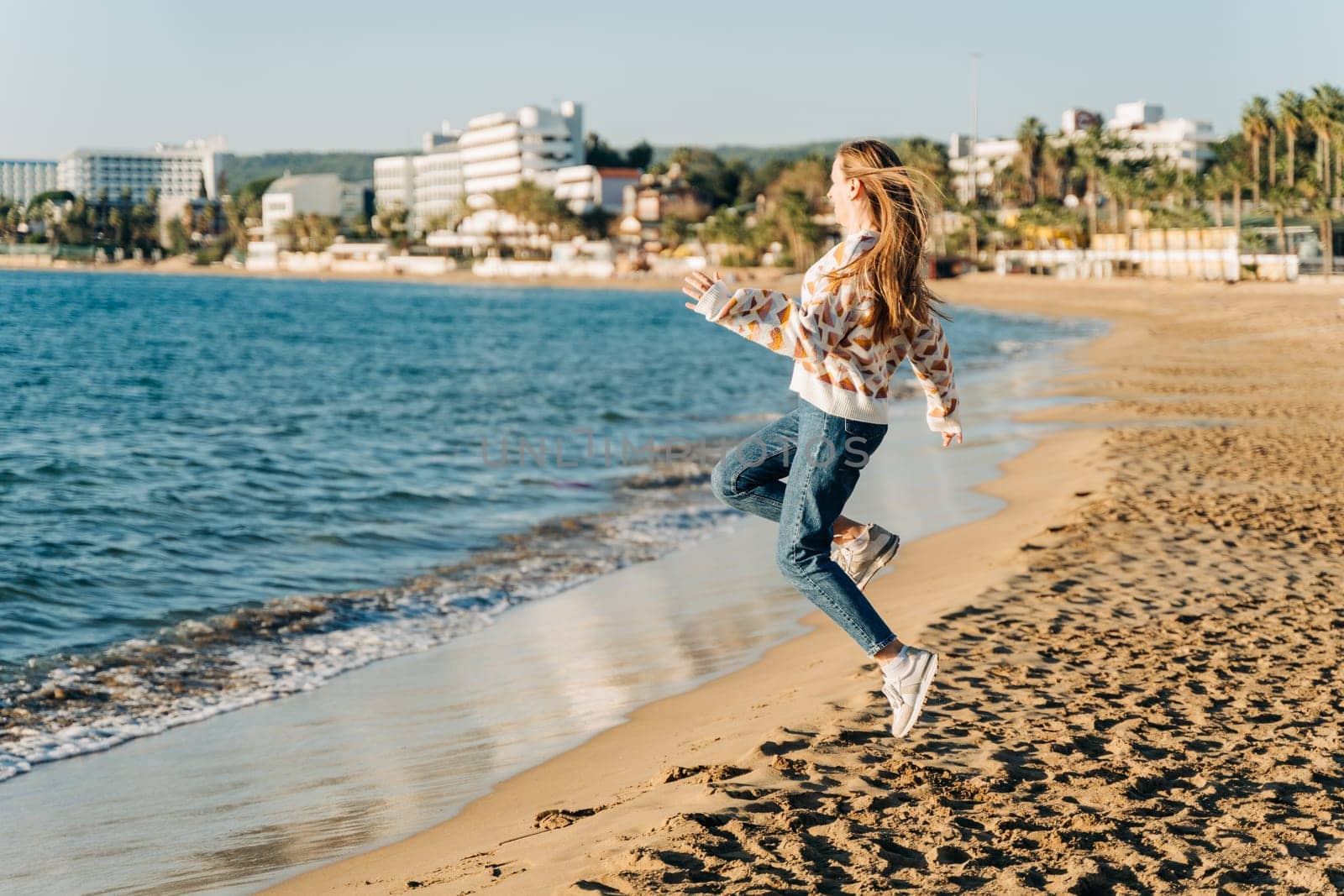 Side portrait of young beautiful woman in cozy sweater running to the winter sea waves. Cute attractive girl having fun jumping near autumn ocean on the beach sand by Ostanina