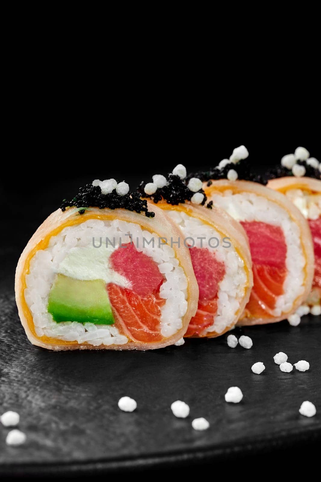 Closeup of delicate sushi rolls wrapped in mamenori filled with raw tuna, salmon, avocado and cream cheese topped with black tobiko and crispy airy rice balls served on slate board. Japanese food