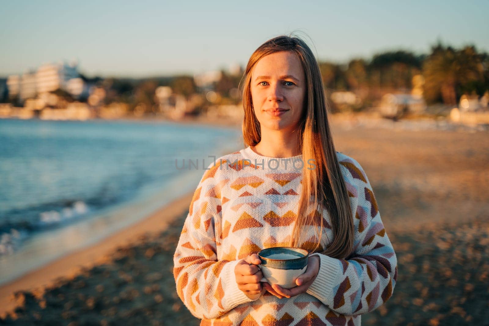 Young beautiful girl in cozy sweater holding coffee cup while enjoying winter sun on seaside shore during mild sunset. Cute attractive woman enjoying her cup of tea while watching autumn ocean waves.