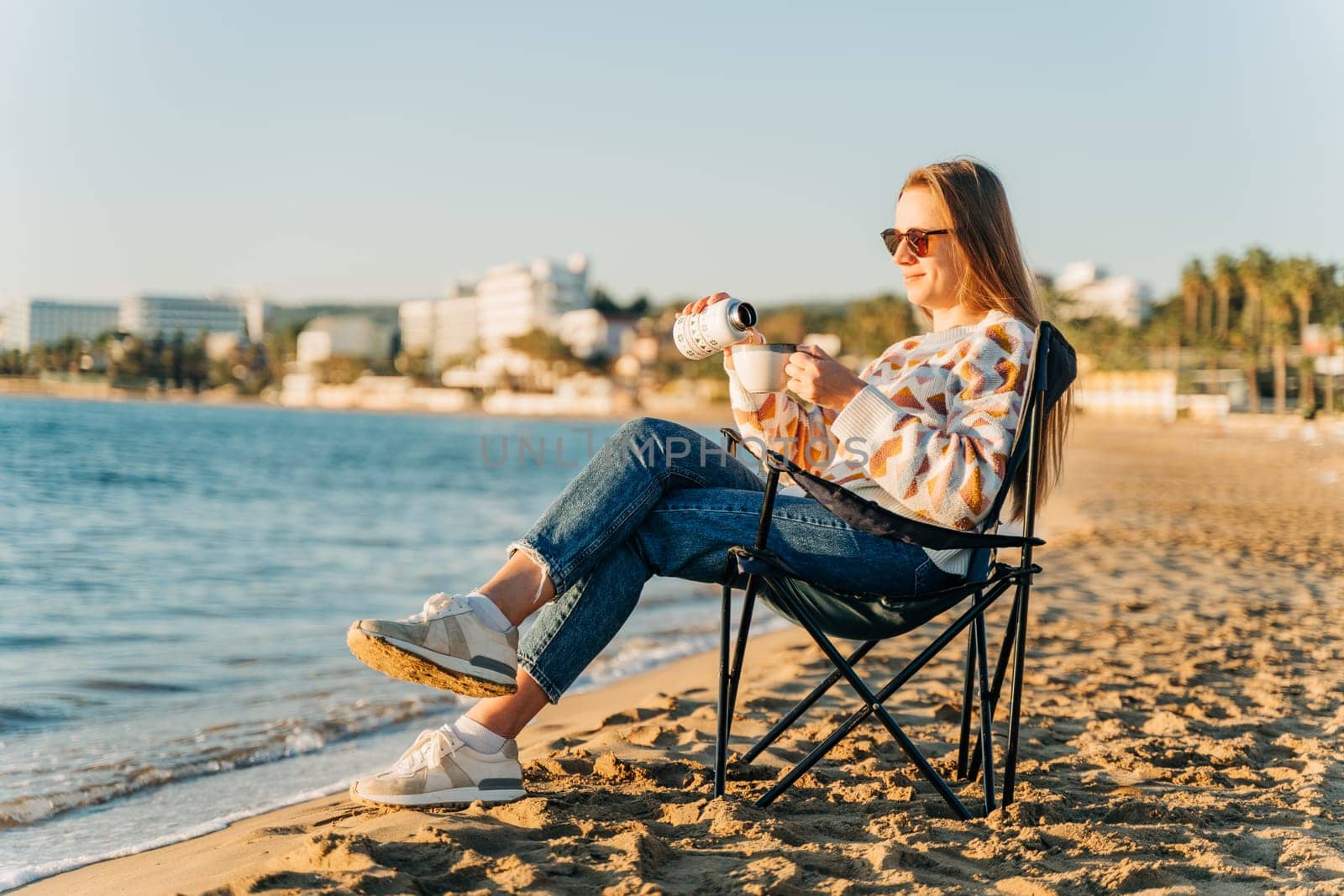 Young beautiful woman in cozy sweater and sunglasses filling cup of coffee from thermos while relaxing on winter seaside sand beach. Cute attractive girl enjoying hot tea taking sunbathe near ocean by Ostanina