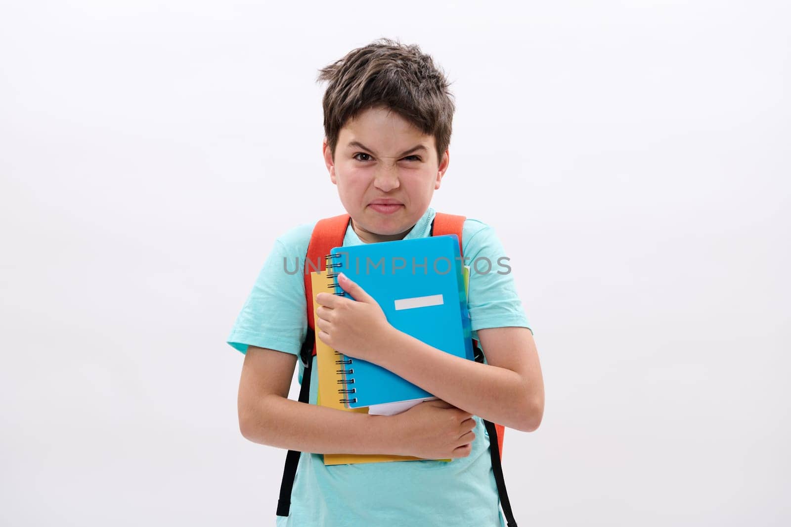 Irritated schoolboy holding workbooks, looking at camera, expressing negative emotions, frustration and anger, feeling unhappy to start a new semester at school, isolated over white studio background