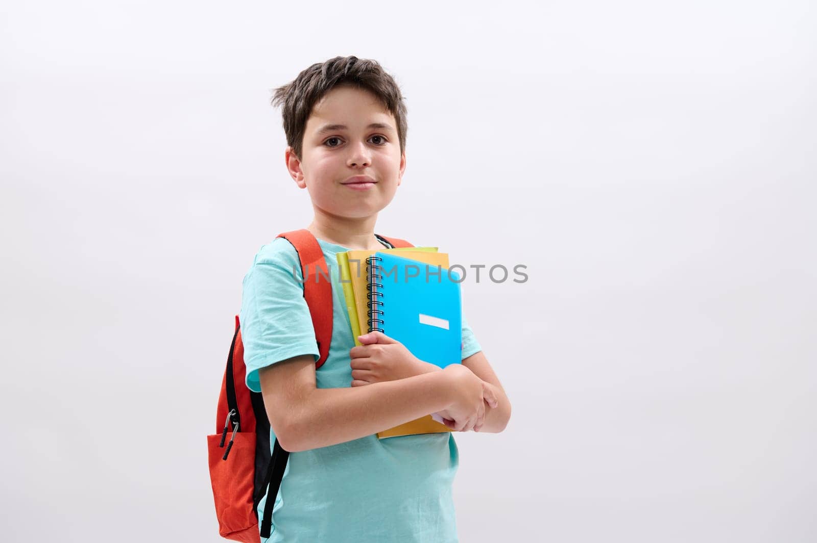 Happy positive teenager smiling looking at camera. Smart schoolboy carrying orange backpack, colorful textbooks and school supplies, isolated on white studio background with copy advertising space