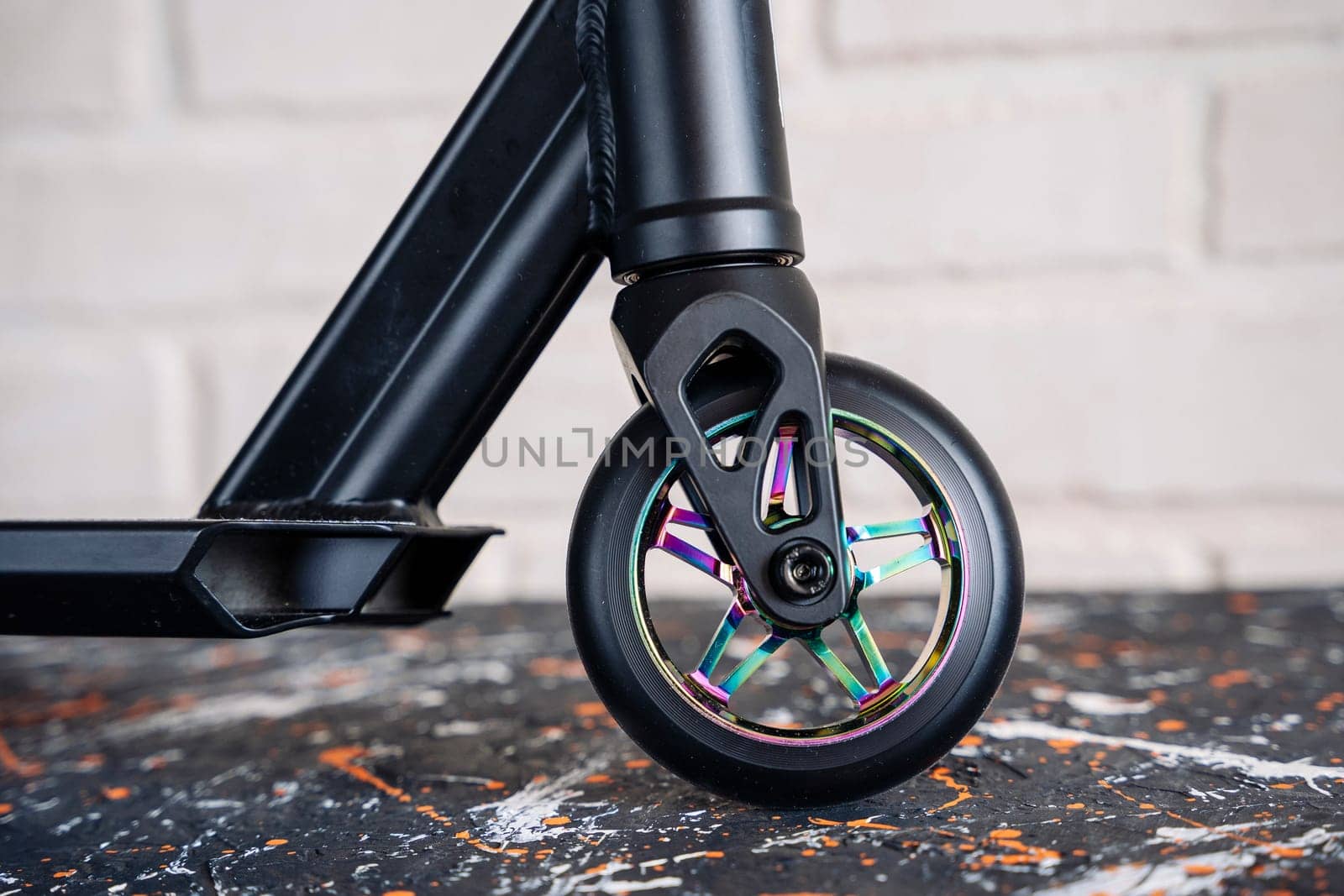 The front wheel of a rainbow-colored stunt scooter against a light brick wall or a durable children's stunt scooter with rubber wheels for extreme skating in a skatepark and on a ramp. Scooter Details