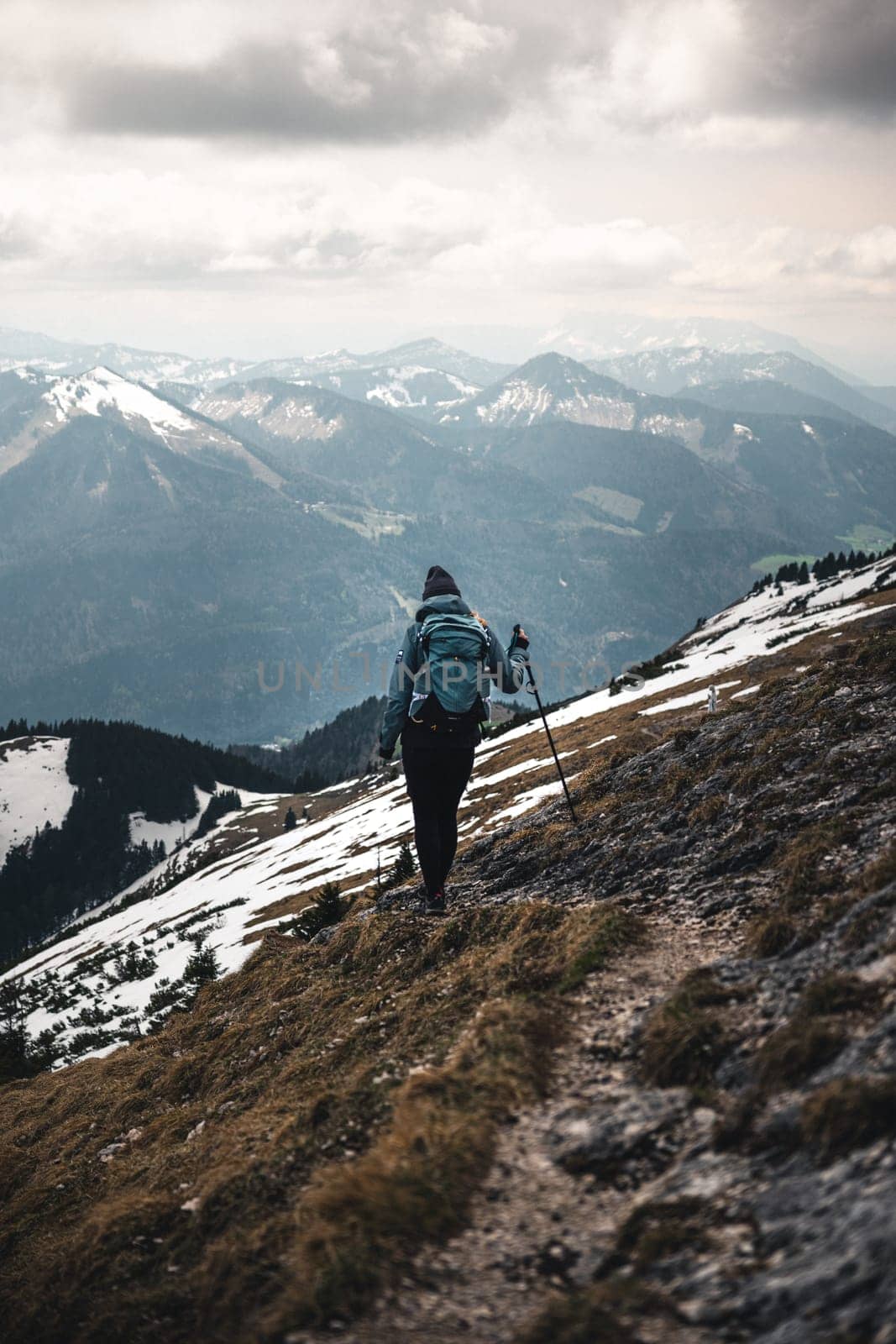 A girl in a blue-gray jacket, dark-gray hat, black leggings, blue-green backpack, and trekking poles is hiking in Schafberg mountain, Innsbruck, Austria. The sky is cloudy, she walks down the path, it's already the end of spring, but the snow is still on the ground.