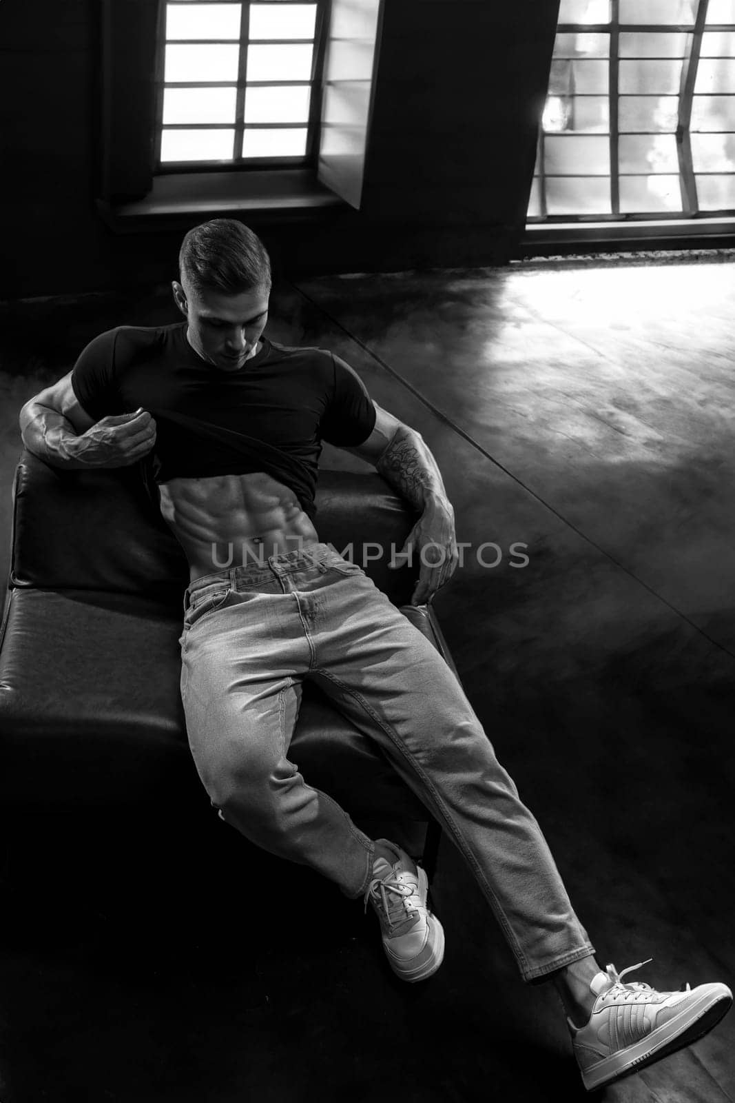 Young sexy male bodybuilder demonstrates perfect abs by lifting his t-shirt up. Handsome athletic fitness model in jeans and t-shirt posing in the studio sitting on a leather sofa against window