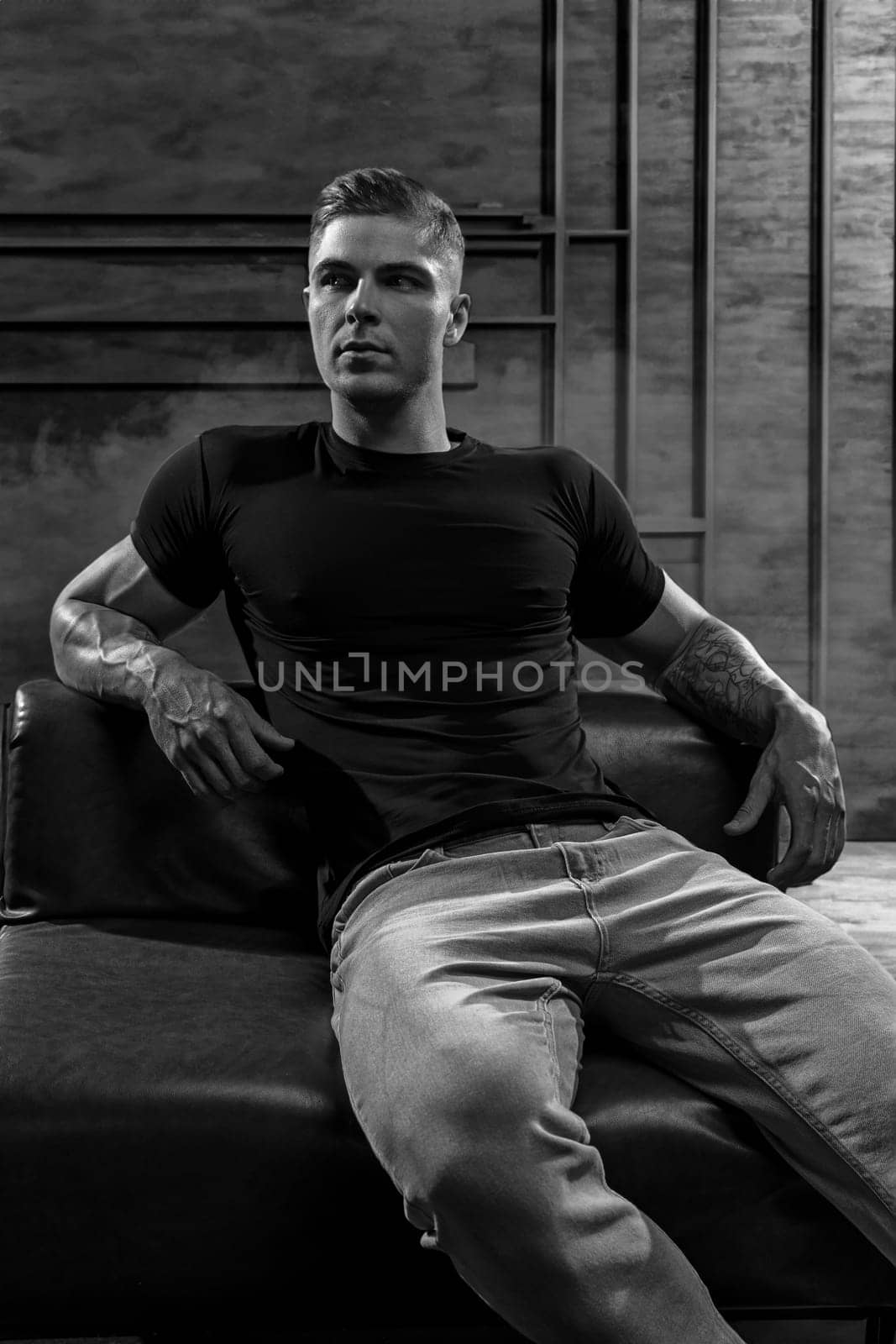 Young sexy bodybuilder athlete posing while sitting on a leather sofa against the background of a stylized loft-style wall. Fitness model in black t-shirt and jeans, black and white photo