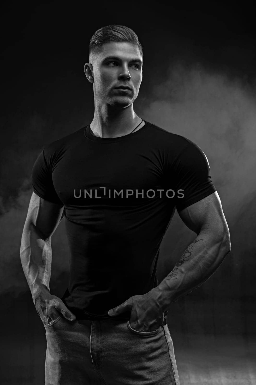 Muscular model sports young man in jeans and black t-shirt on black background. Fashion portrait of brutal sporty healthy strong muscle guy with a modern trendy hairstyle. Black and white photo