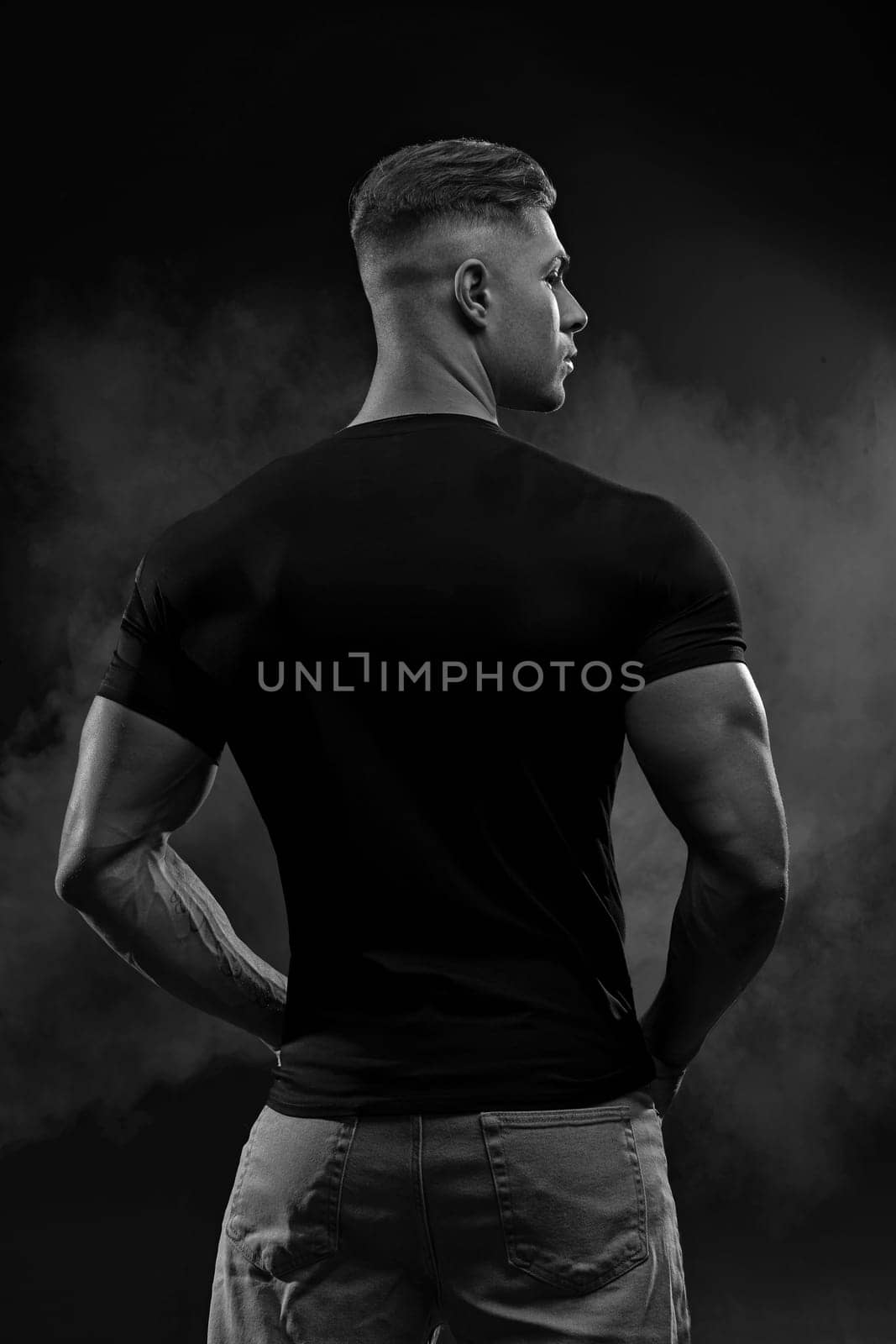 Muscular model sports young man in jeans and black t-shirt on black background back view. Fashion portrait of brutal sporty healthy strong muscle guy. Black and white photo
