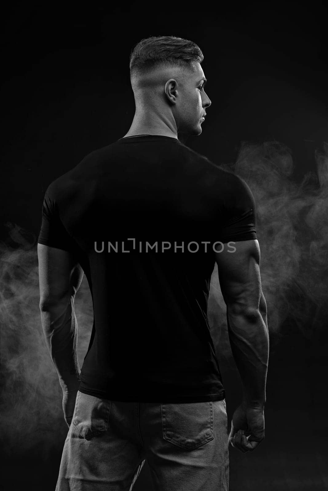 Muscular model sports young man in jeans and black t-shirt on black background back view. Fashion portrait of brutal sporty healthy strong muscle guy black and white photo