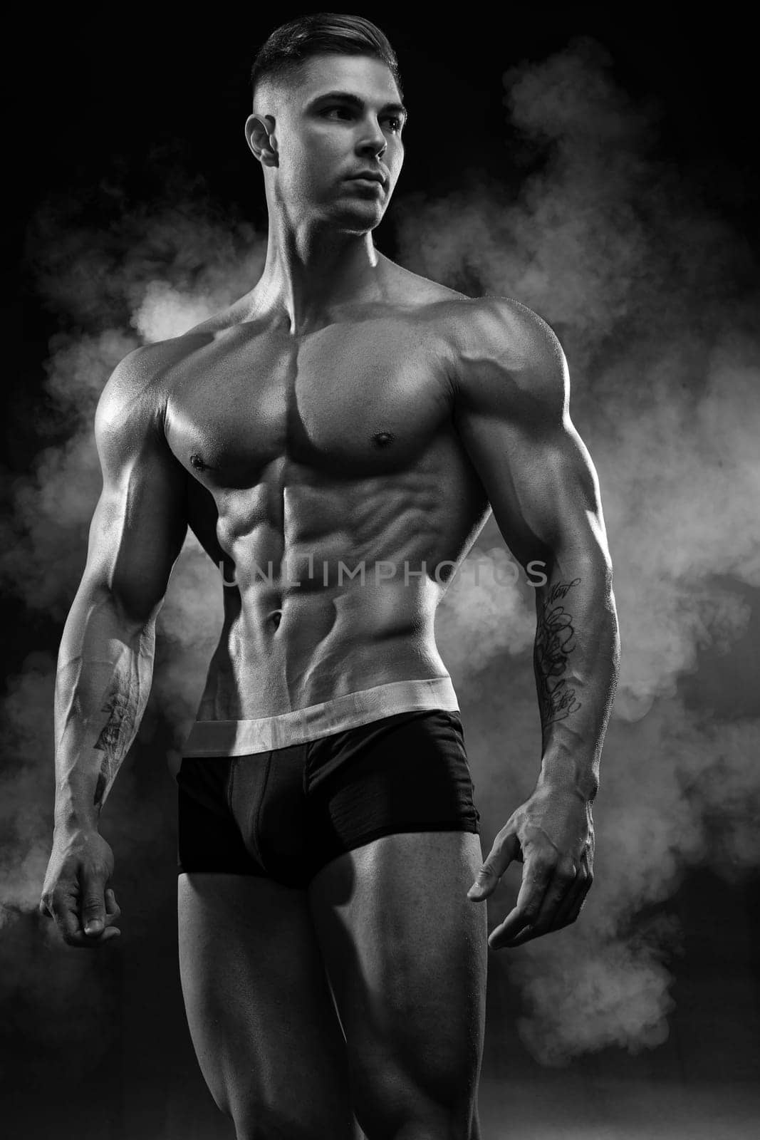 Bodybuilder adult muscular male model posing on black background in shorts demonstrates perfect abs with a low percentage of fat, black and white photo