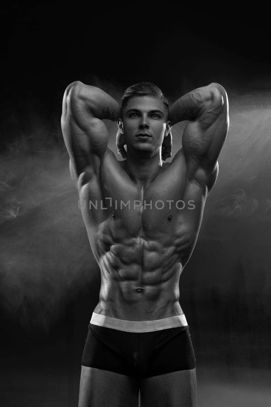 Bodybuilder adult muscular male model posing on empty background in shorts, raising his arms up, demonstrates the latissimus dorsi muscles and perfect abs with low percentage of fat