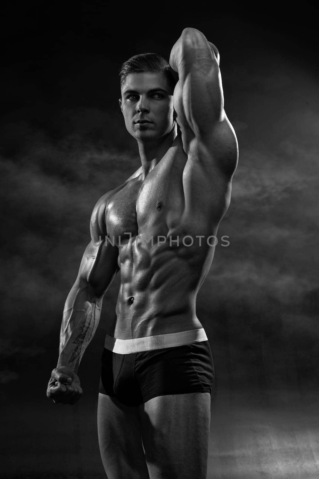 Muscular man shows his perfect low-fat body with six-pack abs on black cyclorama in the studio. Male fitness model macho in shorts with naked torso black and white photo