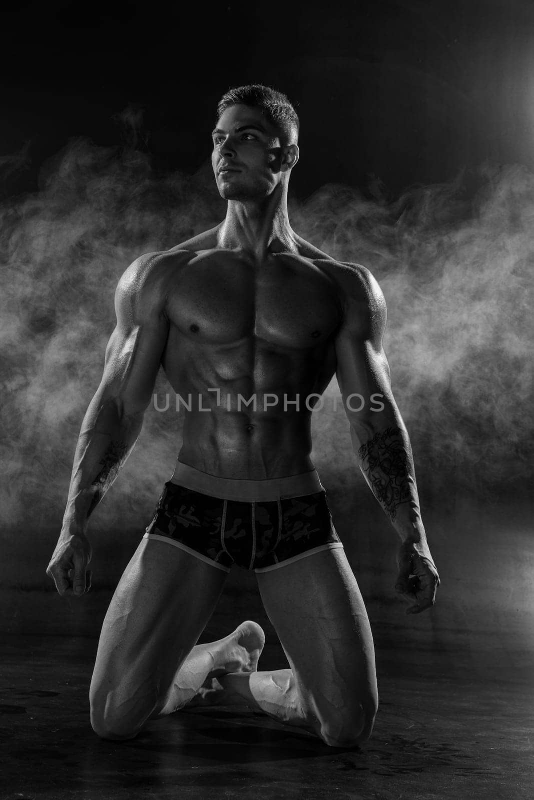 A strong athletic man poses shirtless standing on knees in black studio. Athletic muscular body with six pack abs and low percentage of fat black and white photo