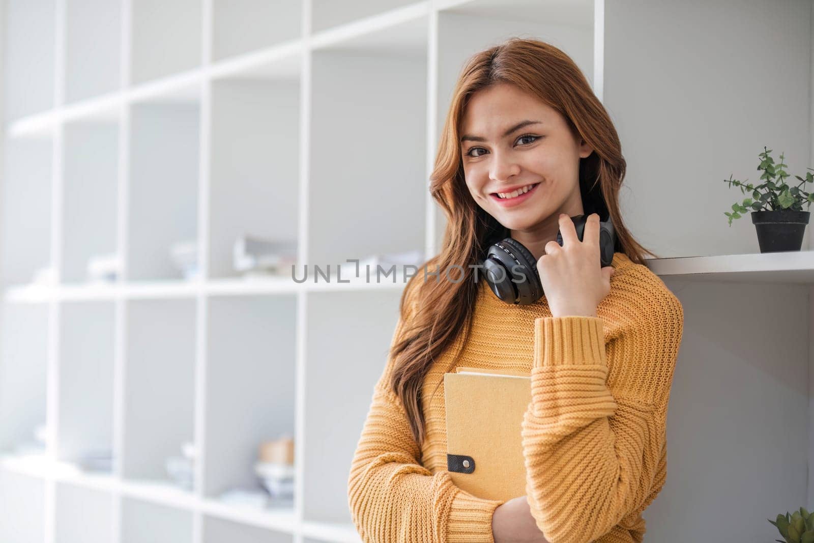 A portrait of an attractive young Asian female in a cozy yellow sweater stands inside a coffee shop with a book and headphones. college student, freelancer by wichayada