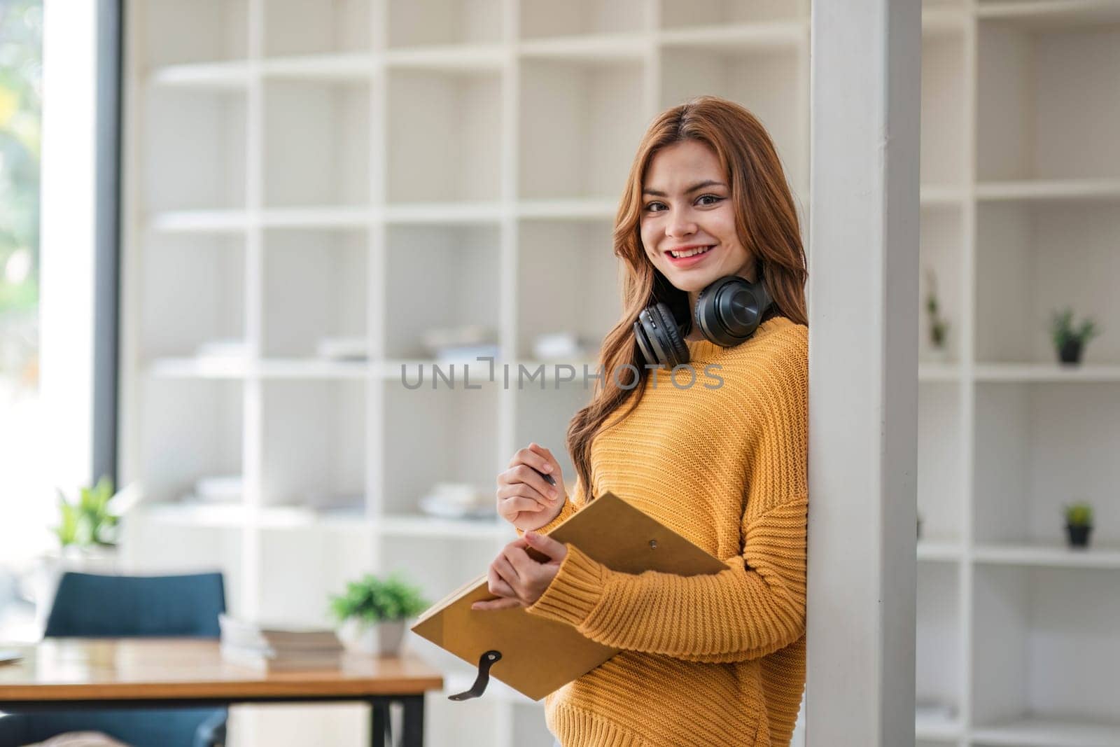 A portrait of an attractive young Asian female in a cozy yellow sweater stands inside a coffee shop with a book and headphones. college student, freelancer by wichayada