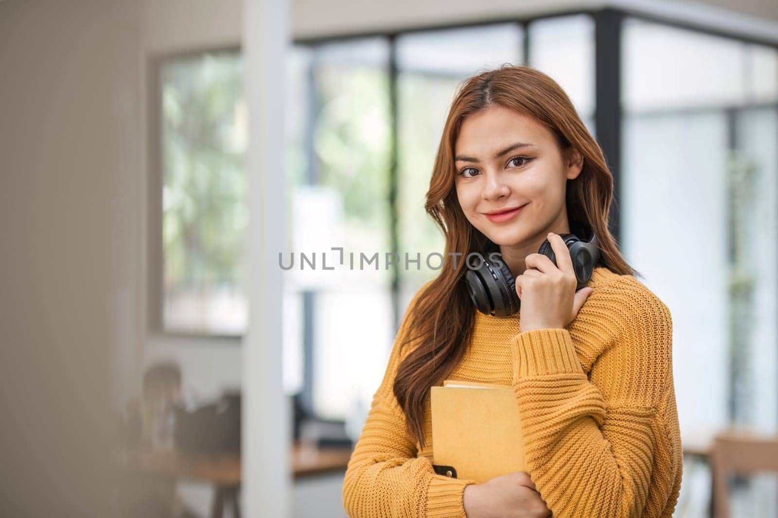 A portrait of an attractive young Asian female in a cozy yellow sweater stands inside a coffee shop with a book and headphones. college student, freelancer by wichayada