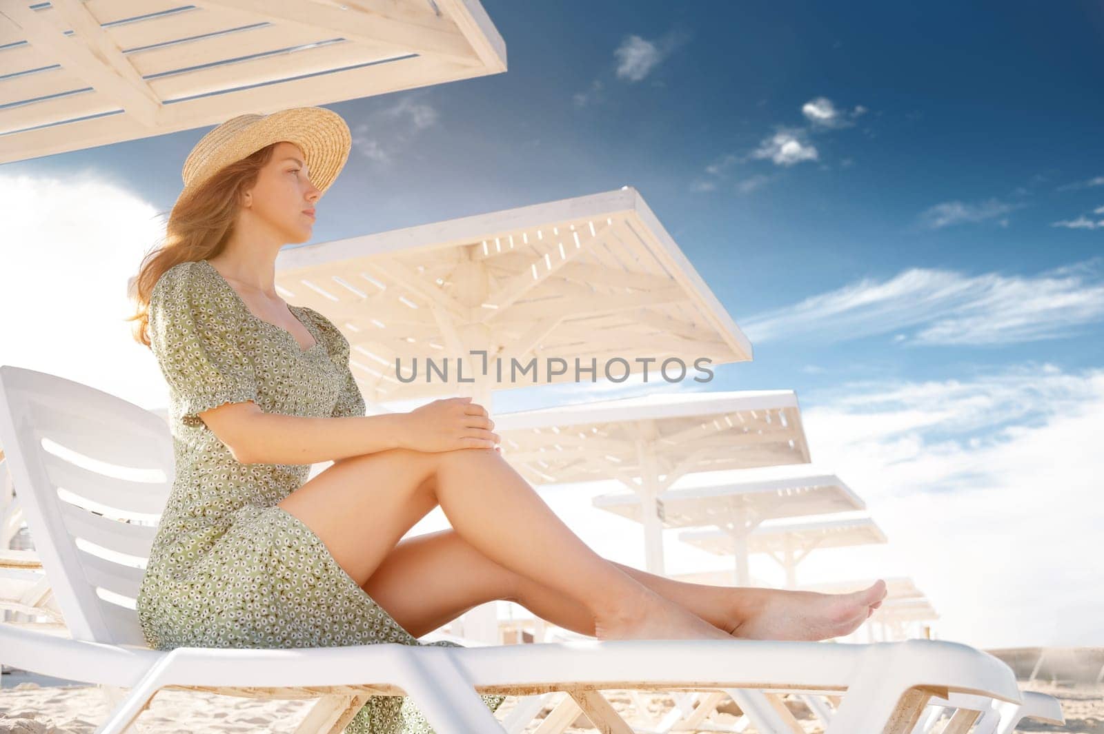 Young attractive caucasian caucasian woman in a straw hat and green dress sitting on a chaise longue. under a wooden umbrella on a sunny day looks at the sea