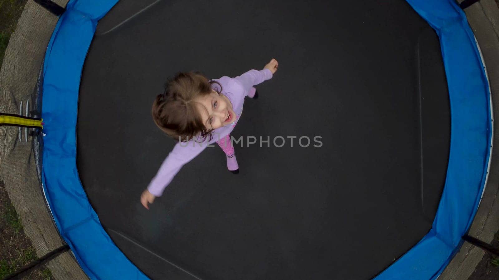 Top view of a jumping girl from a drone. A little girl is jumping on a trampoline in the yard. The camera is spinning. by Puzankov
