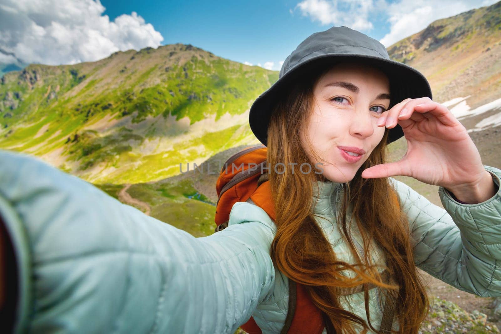 Attractive young caucasian woman in panama, down jacket and backpack taking selfie and half heart gesture high in mountains looking and winking at camera. Invitation to travel in the mountains and trekking.