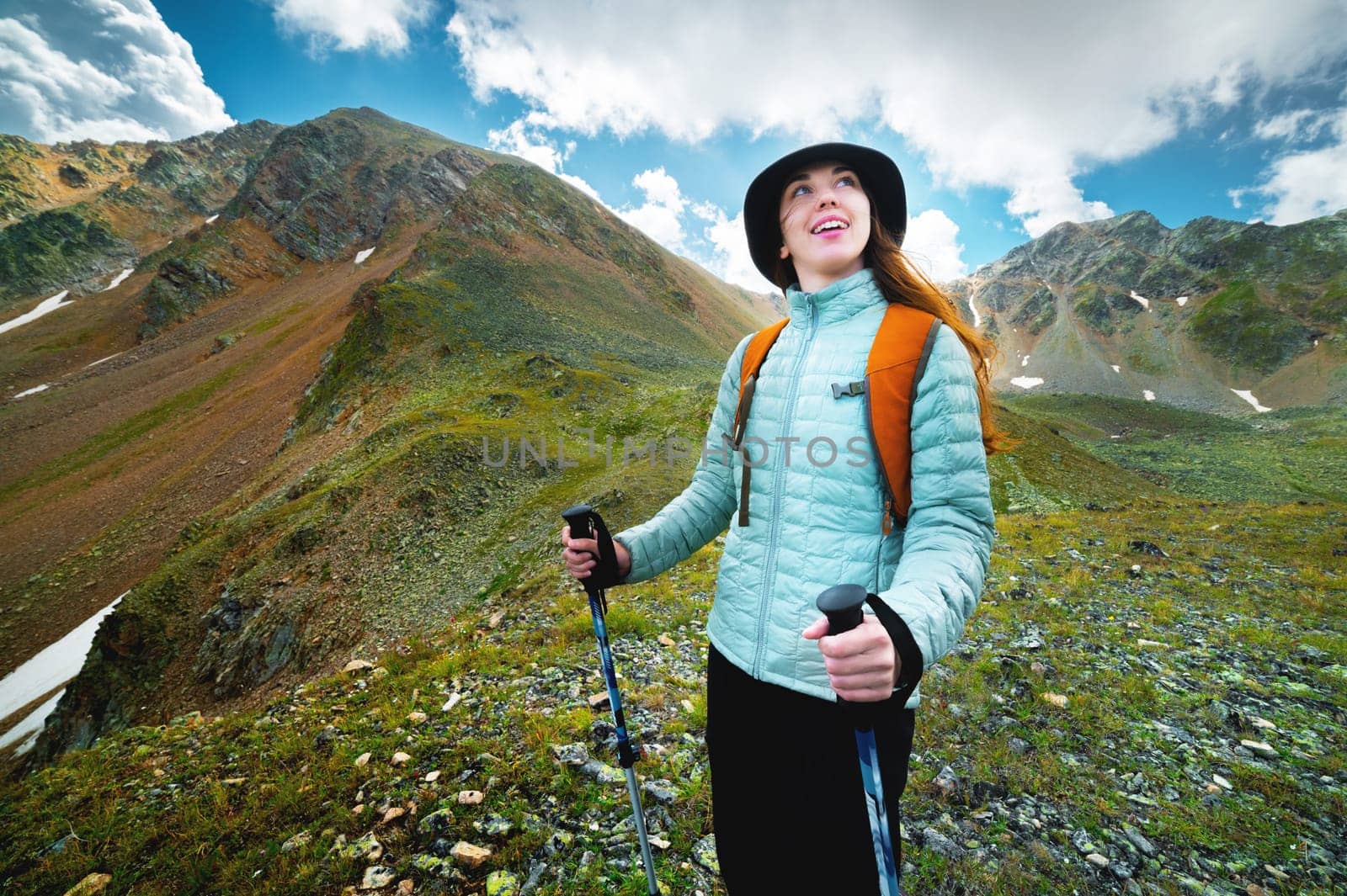 Active young woman standing in a meadow high in the mountains in the afternoon with a trekking stick. The girl holds trekking sticks in her hands and enjoys the views while hiking in the mountains.