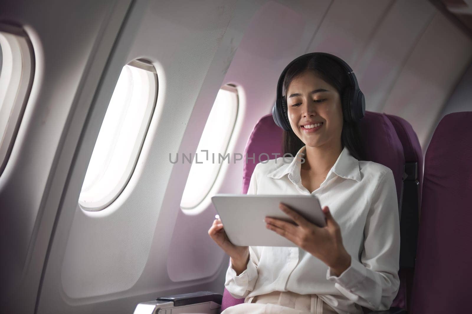 Successful beautiful young asian business woman sits in airplane cabin plane and works on digital tablet with stylus. Flying at first class.
