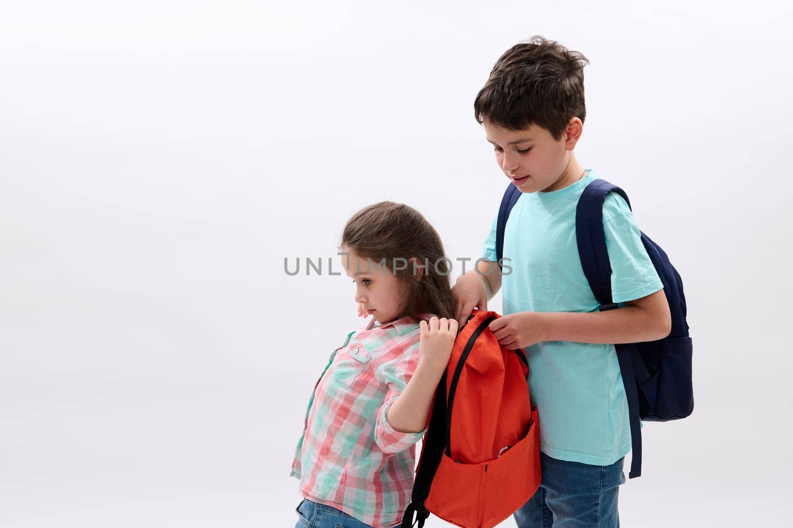 Handsome Caucasian teen boy, a loving caring older brother helps his younger sister to put on a backpack on back, going to school, isolated on white background. Education. New semester. Copy ad space