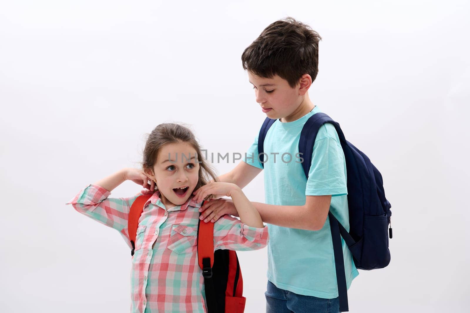 Handsome preteen schoolboy, loving caring brother puts on backpack on his younger sister back, preparing her to the school, isolated on white background. Happy children with school supplies. Education