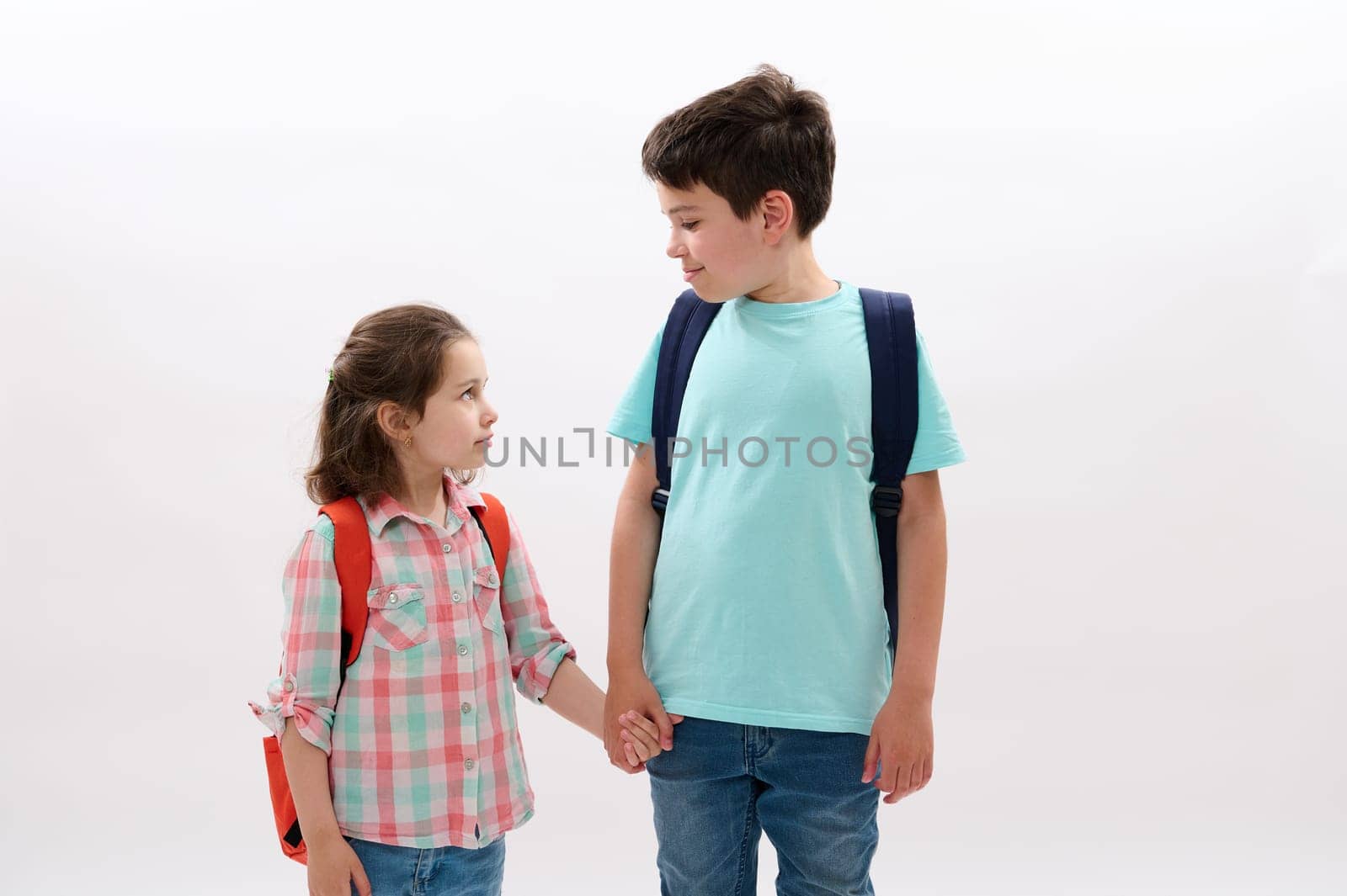 Happy schoolkids, brother and sister holding hands, going to the school, smiling looking at each other, isolated on white backdrop.