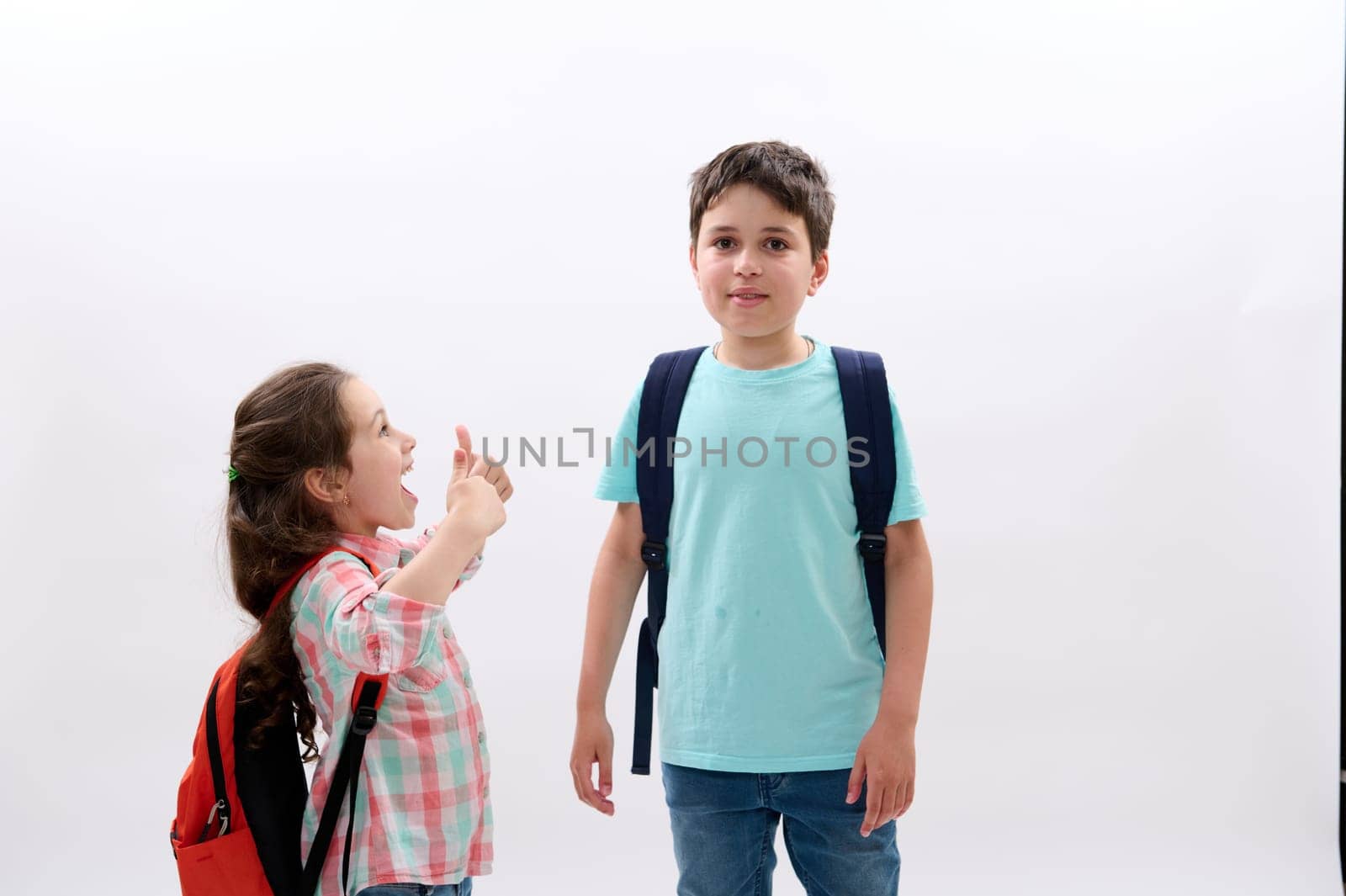 Smart school kids with backpacks, mischievous little girl smiles looking at her older brother, isolated white background by artgf