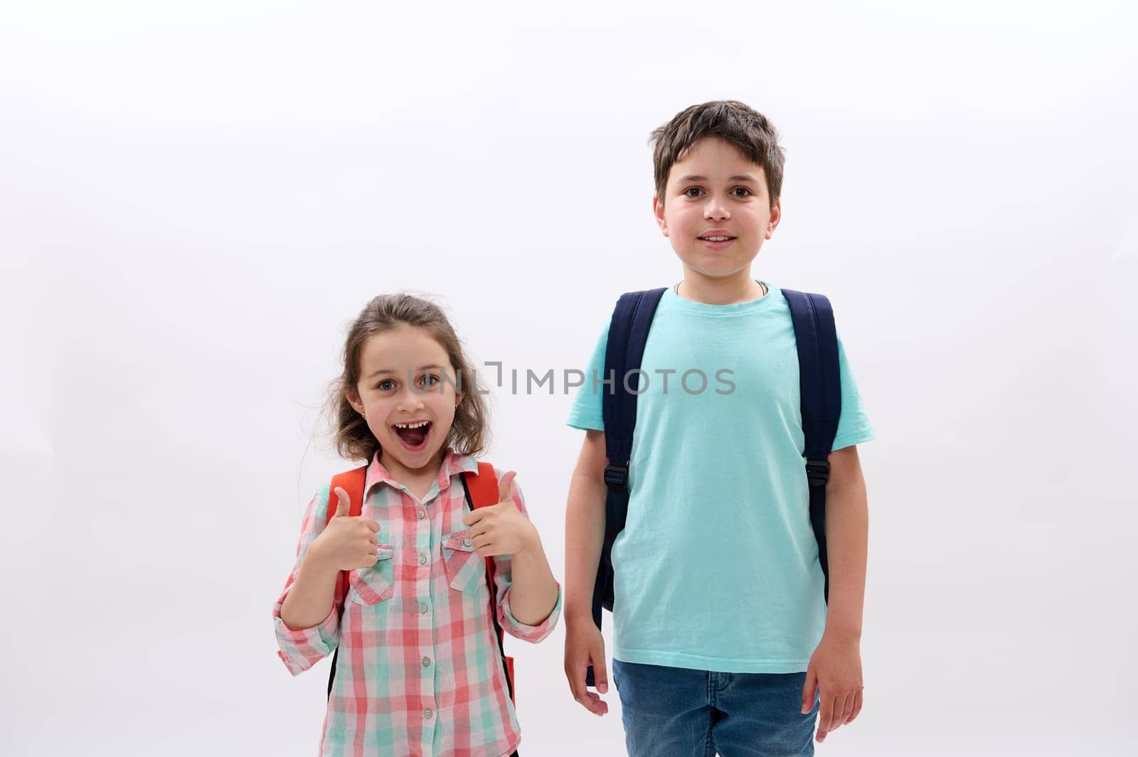 Adorable cheerful school kids - preschooler child girl and teen boy in casual clothes, expressing positive emotions, carrying backpacks, smiling looking at camera, isolated on white studio background