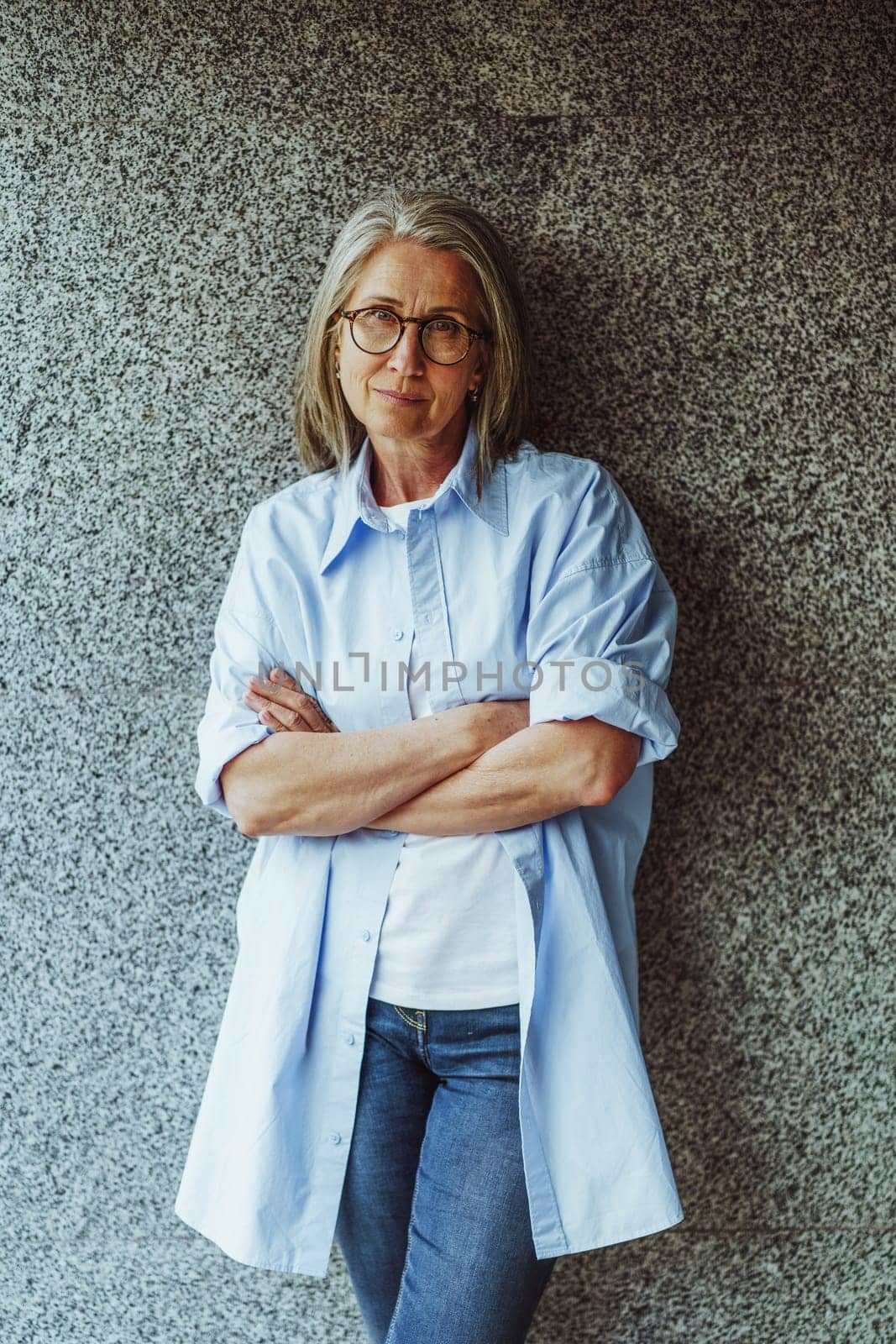 mature lady with gray hair, standing near wall. Lady's confident demeanor, reflecting her wisdom and experience. She exudes air of elegance and sophistication, with stylish appearance and poised posture. High quality photo