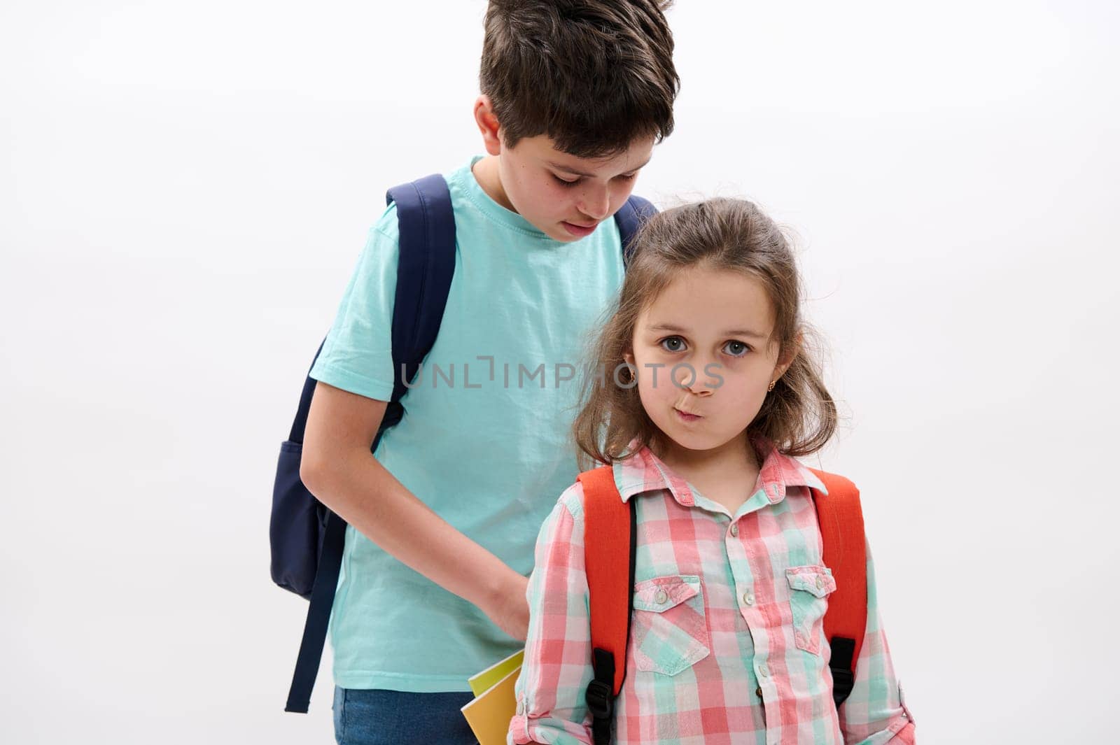Lovely little child girl, mischievous preschooler kid makes faces looking at camera while her older brother puts workbooks inside her backpack, isolated over white background. Back to school concept