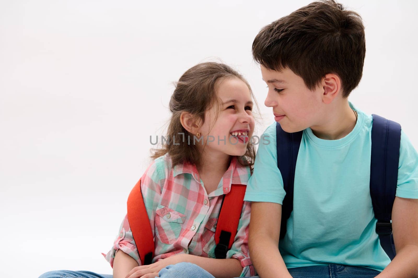 Mischievous little girl, shows tongue and makes faces, smiles to her older brother, isolated over white background. Cheerful smart school Kids with backpacks having fun together. Family relationships